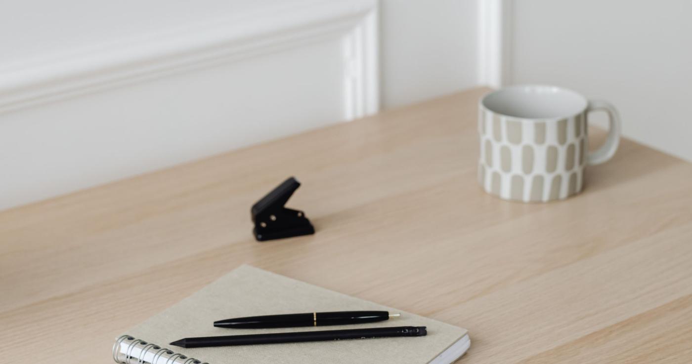 Notizbuch mit Stift und Bleistift auf einem hellen Holztisch, dahinter eine Tasse